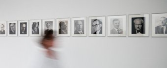 Symbolfoto: Unscharfe Umrisse einer Person, die auf einem Flur an einer Foto-Galerie ehemaliger HRK-Präsidenten vorbeigeht.