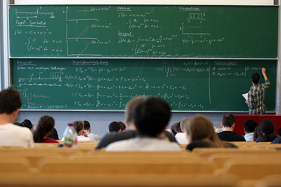 Lehrsituation im Seminar mit einer Lehrperson, die an die Tafel schreibt, während Studierende zuhören.