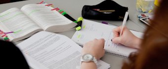 Symbolfoto: Auf einem Tisch liegen aufgeschlagene Lehrbücher. Eine Frau macht sich Notizen.